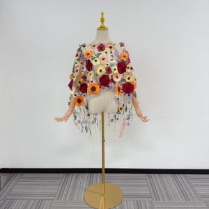 Peut inclure: Une cape transparente ornée de fleurs brodées dans des tons de rouge, orange, jaune et violet. La cape est présentée sur un mannequin avec des bras en bois et un support doré. Le motif floral est détaillé et couvre tout le vêtement.