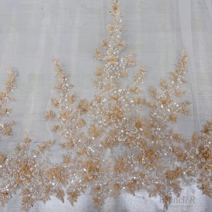 K&ouml;nnte beinhalten: Ein transparenter wei&szlig;er T&uuml;llstoff mit aufwendigen Stickereien in Beige- und Champagnert&ouml;nen. Das Design umfasst dreidimensionale florale Applikationen, zarte Blattmuster und verstreute klare Pailletten, die f&uuml;r einen dezenten Glanz sorgen.