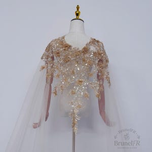 K&ouml;nnte beinhalten: Ein transparentes beiges Cape mit aufwendigen Blumenstickereien, Pailletten und Perlen. Das Kleidungsst&uuml;ck zeigt eine dichte Anordnung aus Stoffblumen und schimmernden Verzierungen, die sich an den Schultern konzentrieren und den R&uuml;cken hinunterlaufen.