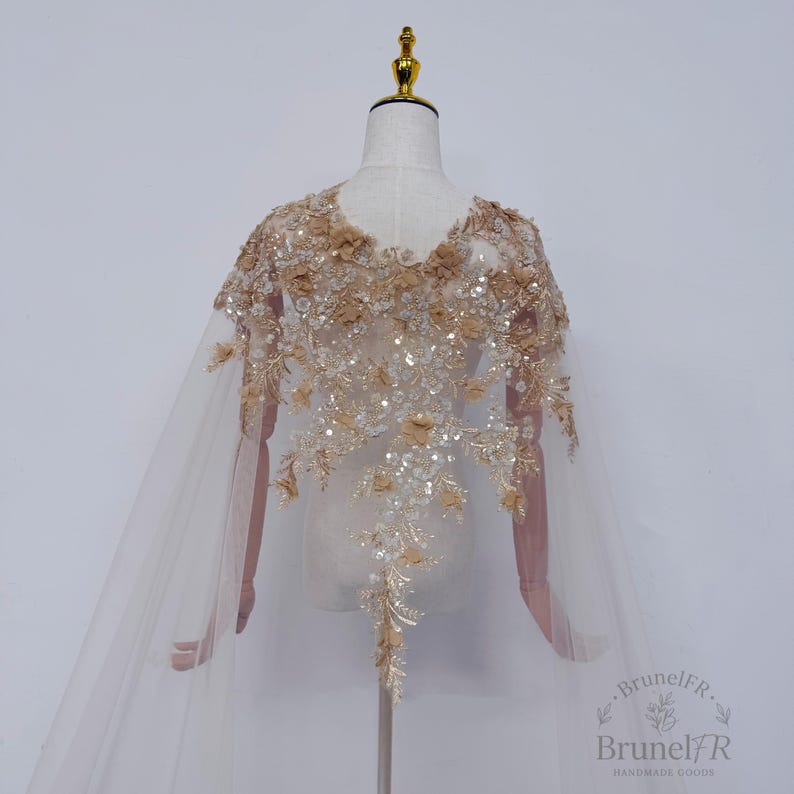 K&ouml;nnte beinhalten: Ein transparentes beiges Cape mit aufwendigen Blumenstickereien, Pailletten und Perlen. Das Kleidungsst&uuml;ck zeigt eine dichte Anordnung aus Stoffblumen und schimmernden Verzierungen, die sich an den Schultern konzentrieren und den R&uuml;cken hinunterlaufen.