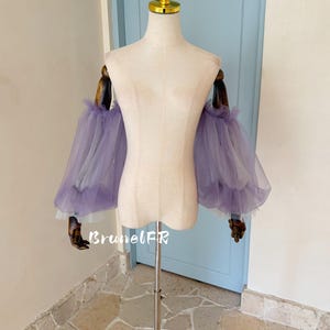 Peut inclure: Un mannequin présente une paire de manches amovibles en tulle violet. Les manches sont superposées avec un bord à volants et une doublure blanche. Le mannequin est sur un support en métal, avec une porte bleu clair en arrière-plan. Le texte "BrunelFR" est visible.