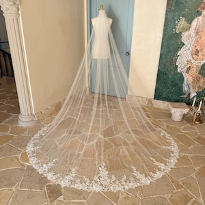 Peut inclure: Un long voile de mariée ivoire recouvrant un mannequin. Le tissu en tulle transparent présente une bordure en dentelle florale complexe le long de l'ourlet arrondi, créant une large traîne qui s'étend sur toute la surface du sol.