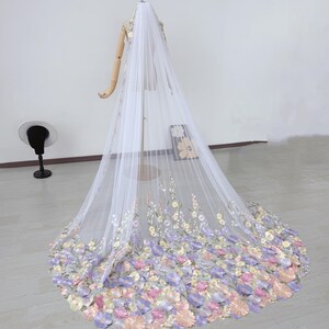 May include: A long, white bridal veil with delicate floral embroidery in pastel colors. The veil is displayed on a mannequin, showcasing the intricate details of the flowers and the flowing fabric. The flowers are in shades of pink, purple, and yellow.