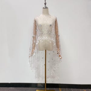 Peut inclure: Une cape transparente blanche ornée de motifs d'étoiles et de feux d'artifice en sequins argentés. Le vêtement présente une encolure ronde, de longs pans fluides et un ourlet festonné décoré de petites étoiles suspendues sur tout le tissu.