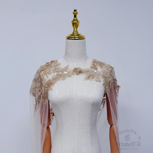 K&ouml;nnte beinhalten: Ein transparentes beiges Capelet mit aufwendiger goldener Blumenstickerei, zarten Perlen und schimmernden Pailletten an Schultern und Ausschnitt. Das Kleidungsst&uuml;ck liegt auf einer Schneiderpuppe und zeigt einen leichten T&uuml;llstoff mit Details.