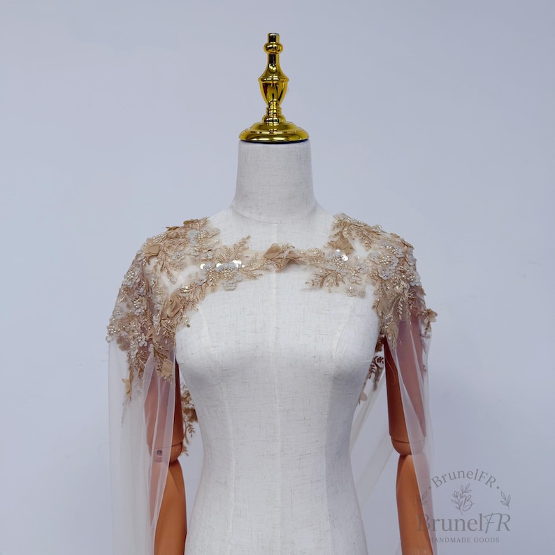 K&ouml;nnte beinhalten: Ein transparentes beiges Capelet mit aufwendiger goldener Blumenstickerei, zarten Perlen und schimmernden Pailletten an Schultern und Ausschnitt. Das Kleidungsst&uuml;ck liegt auf einer Schneiderpuppe und zeigt einen leichten T&uuml;llstoff mit Details.