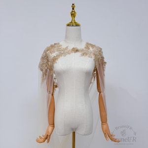 K&ouml;nnte beinhalten: Ein beigefarbenes Spitzen-Cape mit aufwendiger Blumenstickerei, zarten Pailletten und transparentem T&uuml;llstoff. Das Kleidungsst&uuml;ck wird auf einer Schaufensterpuppe pr&auml;sentiert und zeigt einen gewellten Ausschnitt sowie flie&szlig;ende Schulterpartien.