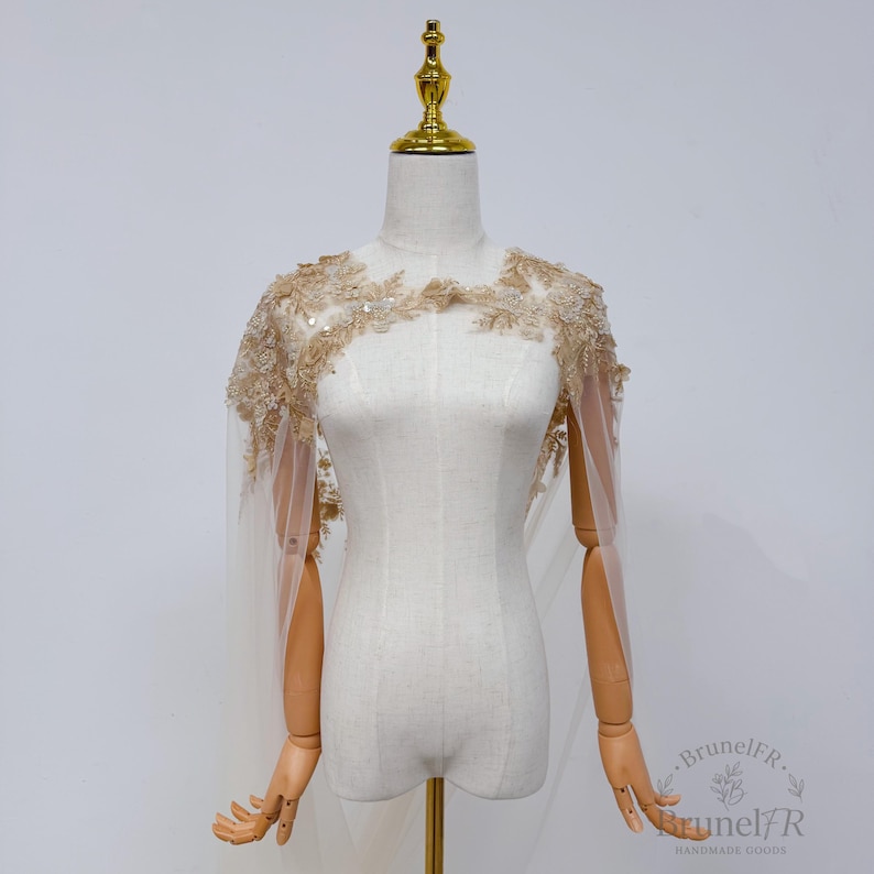 K&ouml;nnte beinhalten: Ein beigefarbenes Spitzen-Cape mit aufwendiger Blumenstickerei, zarten Pailletten und transparentem T&uuml;llstoff. Das Kleidungsst&uuml;ck wird auf einer Schaufensterpuppe pr&auml;sentiert und zeigt einen gewellten Ausschnitt sowie flie&szlig;ende Schulterpartien.