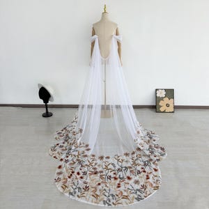 Puede incluir: Una capa de novia de tul blanco con bordados florales en tonos marrón, rojo y verde a lo largo del dobladillo. La capa se exhibe en un maniquí, mostrando su diseño fluido. La capa presenta delicados detalles en los hombros.