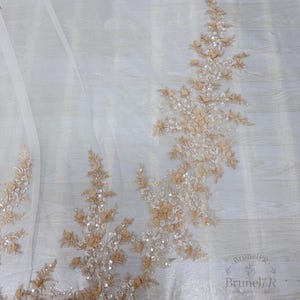 K&ouml;nnte beinhalten: Ein transparenter wei&szlig;er T&uuml;llstoff mit aufwendigen Stickereien in Beige- und Cremet&ouml;nen. Das Design umfasst florale Motive, kleine Stoffbl&uuml;tenbl&auml;tter und verstreute klare Pailletten, die einen strukturierten und schimmernden Effekt erzeugen.