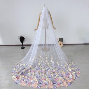 May include: A long, white bridal veil with floral embellishments. The veil is displayed on a mannequin and features a cascade of pastel-colored flowers at the bottom, with delicate embroidered flowers along the edges.