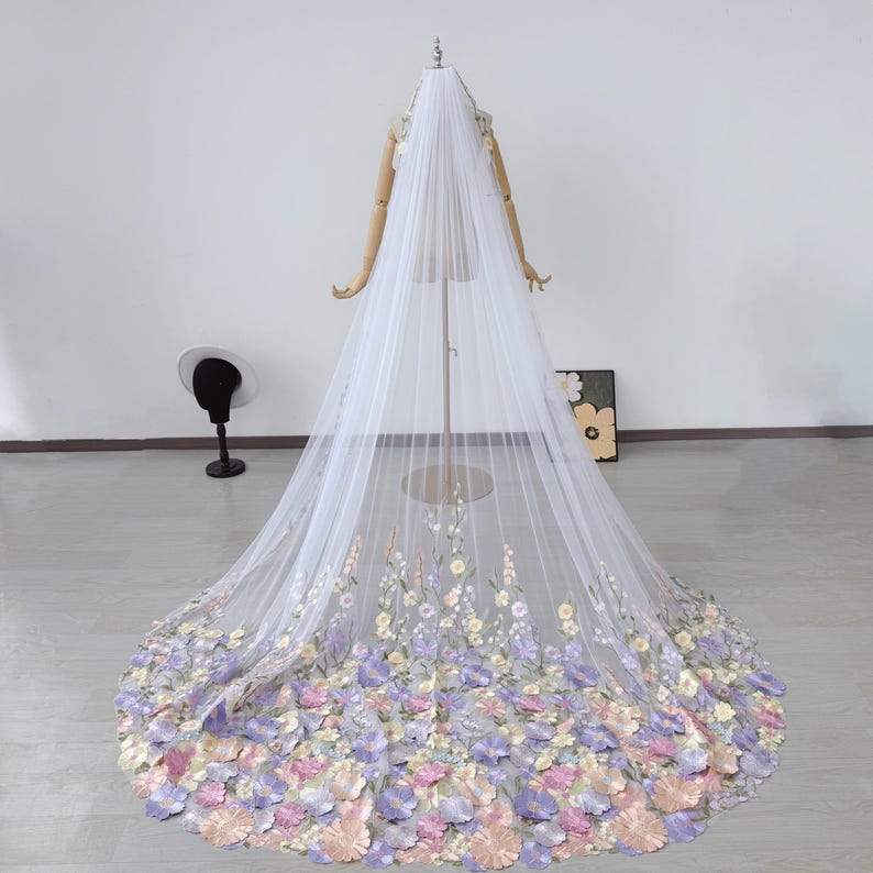 May include: A long, white bridal veil with floral embellishments. The veil is displayed on a mannequin and features a cascade of pastel-colored flowers at the bottom, with delicate embroidered flowers along the edges.