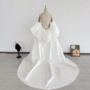 May include: A white bridal gown with a dramatic, oversized ruffled bodice and a flowing skirt. The dress is displayed on a mannequin. A small hat on a stand and a framed floral artwork are in the background.