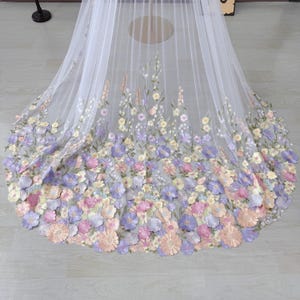 May include: A white tulle fabric with a floral design. The flowers are in shades of pink, purple, and yellow. The flowers are clustered at the bottom of the fabric and fade towards the top.