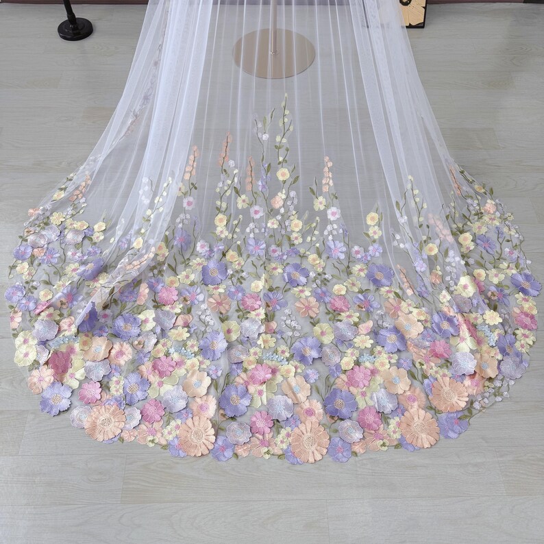 May include: A white tulle fabric with a floral design. The flowers are in shades of pink, purple, and yellow. The flowers are clustered at the bottom of the fabric and fade towards the top.