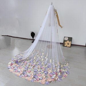 May include: A long, white bridal veil with a floral design. The veil is displayed on a mannequin and features a cascade of pastel-colored flowers in shades of pink, purple, and yellow. The flowers are concentrated at the bottom edge of the veil.
