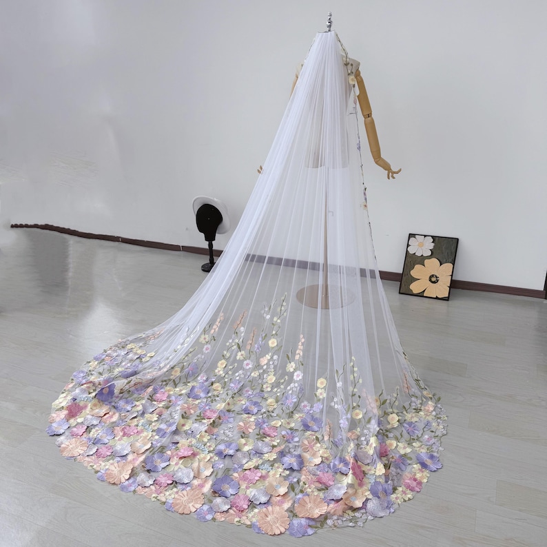 May include: A long, white bridal veil with a floral design. The veil is displayed on a mannequin and features a cascade of pastel-colored flowers in shades of pink, purple, and yellow. The flowers are concentrated at the bottom edge of the veil.