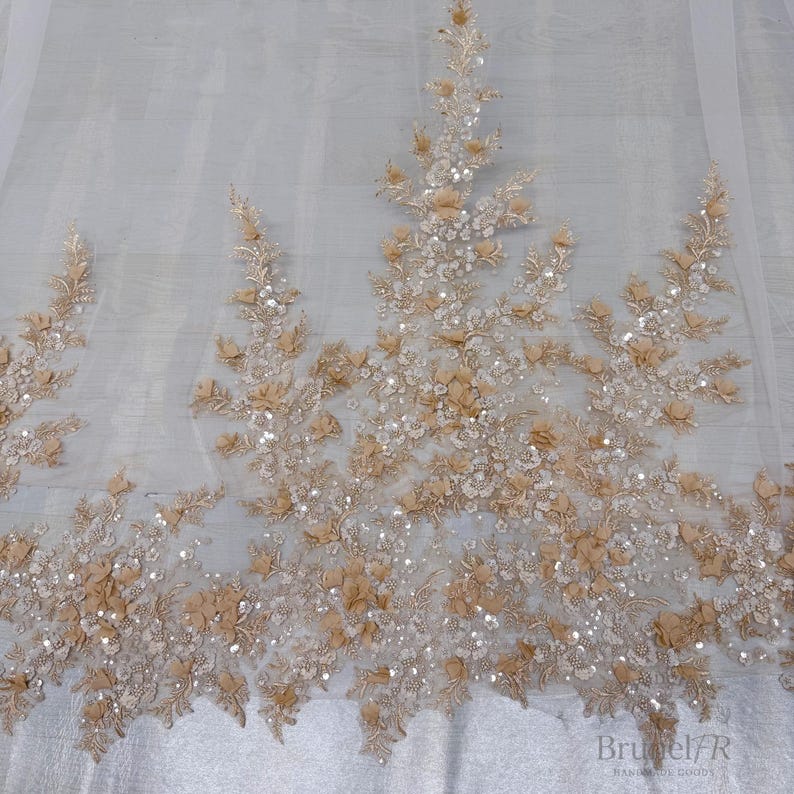 K&ouml;nnte beinhalten: Ein transparenter wei&szlig;er T&uuml;llstoff mit aufwendigen Stickereien in einem hellen Beigeton. Das Design umfasst dreidimensionale florale Applikationen, zarte Blattmuster und verstreute silberne Pailletten, die dem Textil einen dezenten Glanz verleihen.