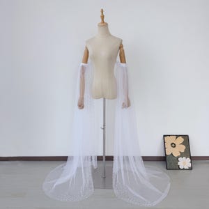 Peut inclure: Une cape de mariée blanche avec de longues manches transparentes et une traîne fluide. La cape est ornée de petites perles éparpillées sur les bords, créant un look délicat et élégant. La cape est présentée sur un mannequin.