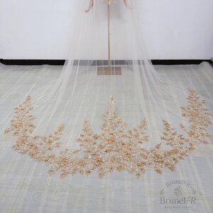 K&ouml;nnte beinhalten: Ein langer, transparenter Brautschleier aus elfenbeinfarbenem T&uuml;ll mit einem aufwendigen, bogenf&ouml;rmigen Saum, der mit filigraner goldener Blumenstickerei, schimmernden Pailletten und kleinen Perlen in einem dichten Muster verziert ist.