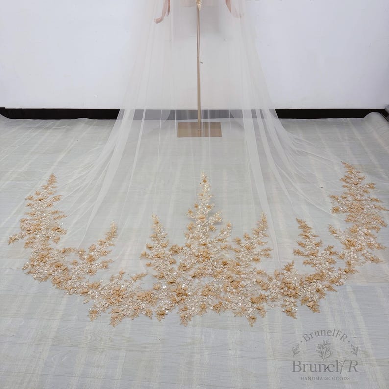 K&ouml;nnte beinhalten: Ein langer, transparenter Brautschleier aus elfenbeinfarbenem T&uuml;ll mit einem aufwendigen, bogenf&ouml;rmigen Saum, der mit filigraner goldener Blumenstickerei, schimmernden Pailletten und kleinen Perlen in einem dichten Muster verziert ist.
