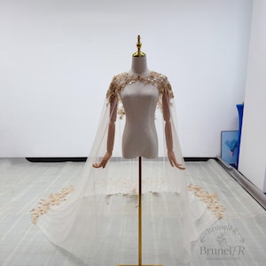 K&ouml;nnte beinhalten: Ein bodenlanger, transparenter Umhang in hellem Cremewei&szlig; mit aufwendigen goldenen Blumenstickereien und Pailletten an Schultern und Ausschnitt. Der Umhang f&auml;llt von den Schultern herab und endet in einer langen Schleppe mit goldenen Verzierungen.