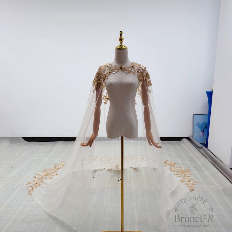 K&ouml;nnte beinhalten: Ein bodenlanger, transparenter Umhang in hellem Cremewei&szlig; mit aufwendigen goldenen Blumenstickereien und Pailletten an Schultern und Ausschnitt. Der Umhang f&auml;llt von den Schultern herab und endet in einer langen Schleppe mit goldenen Verzierungen.
