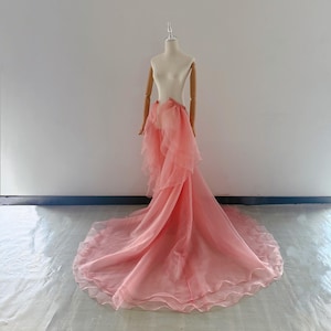 Peut inclure: Une jupe longue jusqu'au sol de couleur pêche douce, confectionnée en organza transparent superposé. Le vêtement présente une taille haute avec des panneaux à volants en cascade créant une silhouette volumineuse et une longue traîne.