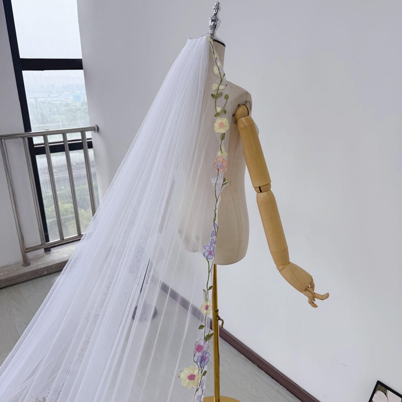 May include: A white bridal veil with a floral embroidered design. The veil is long and flowing, with a delicate, sheer fabric. The floral embroidery features pastel-colored flowers and green leaves, adding a touch of elegance to the wedding accessory.