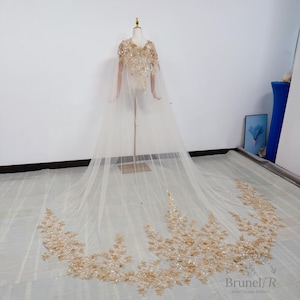 K&ouml;nnte beinhalten: Ein bodenlanger, transparenter Umhang in hellem Beige mit aufwendigen goldenen Pailletten- und Perlenverzierungen an Schultern und Oberteil sowie einem weiten, ausladenden Saum, der mit dichten goldenen Blumenstickereien und Pailletten besetzt ist.