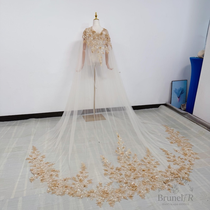 K&ouml;nnte beinhalten: Ein bodenlanger, transparenter Umhang in hellem Beige mit aufwendigen goldenen Pailletten- und Perlenverzierungen an Schultern und Oberteil sowie einem weiten, ausladenden Saum, der mit dichten goldenen Blumenstickereien und Pailletten besetzt ist.