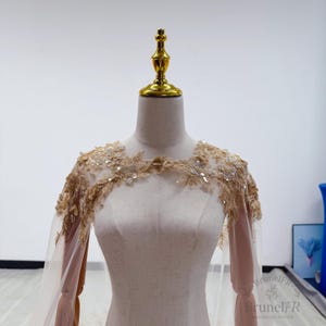 K&ouml;nnte beinhalten: Ein beigefarbenes, transparentes Capelet mit aufwendiger goldener Blumen-Spitzenstickerei an den Schultern und der oberen Brust. Die zarte Spitze ist mit kleinen, schimmernden Pailletten verziert und geht in lange, flie&szlig;ende &Auml;rmel &uuml;ber.