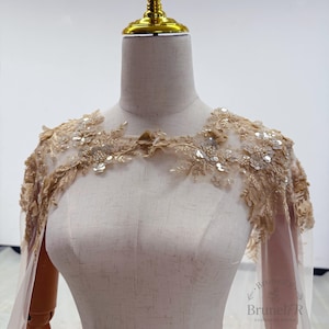 K&ouml;nnte beinhalten: Ein beigefarbener Schulter&uuml;berwurf aus T&uuml;ll mit aufwendiger Goldstickerei, floralen Applikationen und verstreuten Pailletten. Das Kleidungsst&uuml;ck wird auf einer Schneiderpuppe pr&auml;sentiert und umfasst transparente Stoffbahnen, die von den Schultern herabh&auml;ngen.