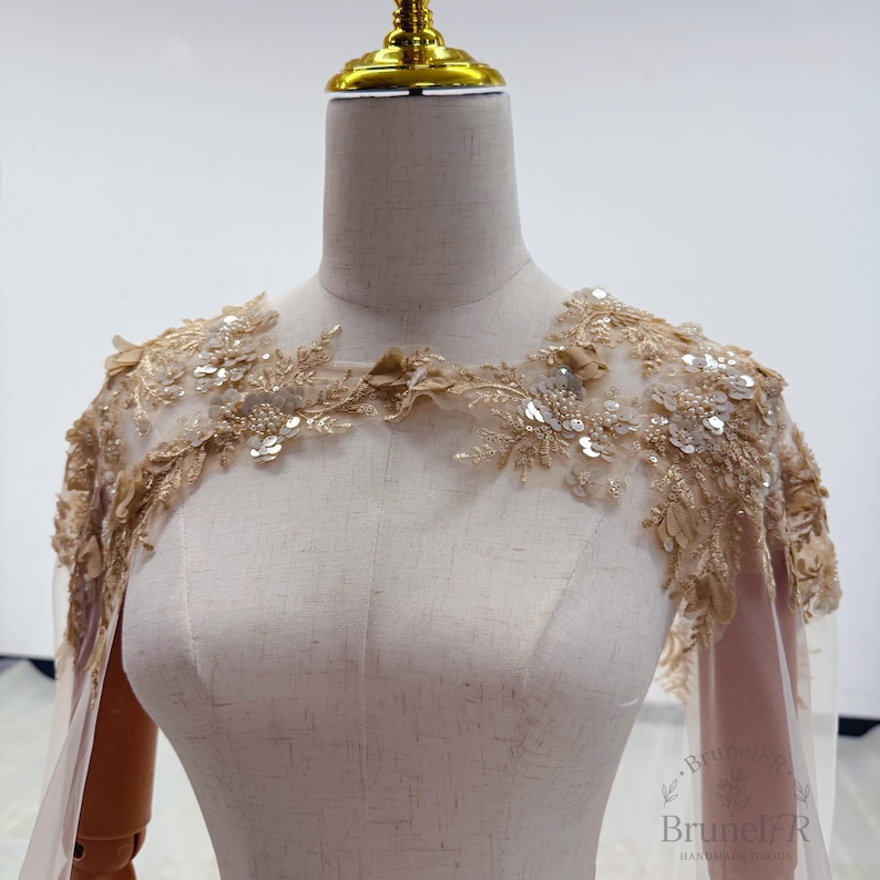 K&ouml;nnte beinhalten: Ein beigefarbener Schulter&uuml;berwurf aus T&uuml;ll mit aufwendiger Goldstickerei, floralen Applikationen und verstreuten Pailletten. Das Kleidungsst&uuml;ck wird auf einer Schneiderpuppe pr&auml;sentiert und umfasst transparente Stoffbahnen, die von den Schultern herabh&auml;ngen.
