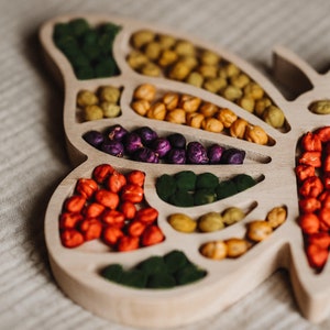 Könnte beinhalten: Ein hölzernes, schmetterlingsförmiges Tablett gefüllt mit bunten Gegenständen. Das Tablett ist in Abschnitte unterteilt, die jeweils eine andere Farbe kleiner Objekte enthalten, darunter Grün, Gelb, Orange und Lila. Das Tablett liegt auf einem neutralfarbenen Stoff.