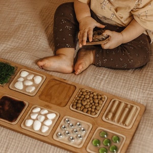 Puede incluir: Un tablero sensorial de madera con varios compartimentos llenos de diferentes texturas y objetos, incluyendo hierba verde, conchas blancas, granos de café marrones, arena marrón, piedras blancas, cuentas de metal plateadas y cuentas de plástico verdes. Un niño está jugando con el tablero.