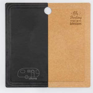 May include: A two-toned cutting board with a black and tan color scheme. The black side features a white outline of a camper and the word "ellhome". The tan side has the text "Oh Darling - let's be - Adventures". A hole is at the top.