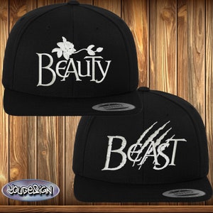 Könnte beinhalten: Zwei schwarze Baseballkappen mit weißer Stickerei. Eine Kappe trägt die Aufschrift "Beauty" mit einem Blumendesign. Die andere Kappe trägt die Aufschrift "Beast" mit Krallenabdrücken.