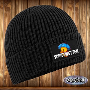 Könnte beinhalten: Schwarze, gerippte Strickmütze mit einem rechteckigen Aufnäher mit einer Cartoon-Ente unter einem blauen Regenschirm. Der Text "SCHIETWETTER" steht darunter. Ein Winteraccessoire.