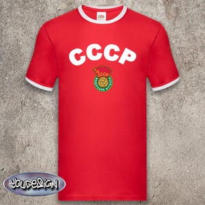 Peut inclure: T-shirt rouge avec bordure blanche au col et aux manches. Le t-shirt affiche le texte blanc "CCCP" au-dessus d'un écusson circulaire avec le texte "CCCP" et un ballon de football. Style vintage.
