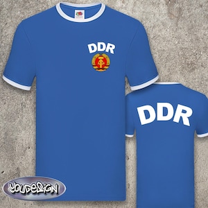 Könnte beinhalten: Zwei blaue T-Shirts mit weißem Besatz und dem Text "DDR" in Weiß. Ein Shirt hat ein rotes und goldenes Emblem mit Hammer und Kompass.