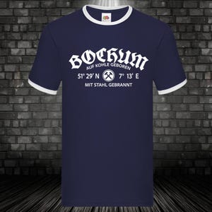 Könnte beinhalten: Marineblaues Ringer-T-Shirt mit weißem Besatz. Das Shirt zeigt das Wort "Bochum" in einer stilisierten Schriftart, zusammen mit Text und Koordinaten. Der Text enthält "AUF KOHLE GEBOREN" und "MIT STAHL GEBRANNT".