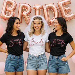 Puede incluir: Tres personas con camisetas y pantalones cortos de mezclilla posan frente a un fondo rosa con una pancarta de globos que dice "BRIDE". La persona central lleva una camisa blanca con "Bride" en purpurina rosa, mientras que las otras llevan camisetas negras con "Team Bride".