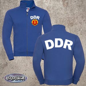 Könnte beinhalten: Blaue Reißverschlussjacke mit den weißen Buchstaben "DDR" auf der Vorder- und Rückseite. Auf der Vorderseite der Jacke befindet sich ein rundes Emblem mit denselben Buchstaben und einem Hammer- und Zirkel-Design. Die Jacke hat einen hohen Kragen.