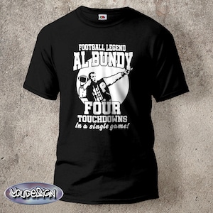 Peut inclure: T-shirt noir avec un graphisme blanc représentant un joueur de football américain et le texte "FOOTBALL LEGEND AL BUNDY FOUR TOUCHDOWNS In a single game!". Le design comprend un ballon et un joueur pointant.