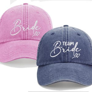 Könnte beinhalten: Zwei Baseballkappen, eine rosa und eine blaue, mit den Wörtern "Bride" und "Team Bride" in weißer Schrift. Jede Kappe hat einen gebogenen Schirm und einen verstellbaren Riemen. Die rosa Kappe hat das Wort "Bride" und ein Herz. Die blaue Kappe hat die Wörter "Team Bride" und ein Herz.