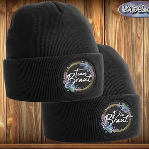 Può includere: Due berretti neri a maglia con risvolto. Ogni berretto presenta una toppa circolare con le parole "Team Braut" e "Die Braut" in corsivo bianco, circondate da un anello dorato e dettagli floreali pastello. Sfondo in legno.