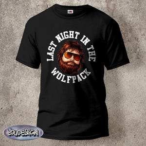 Könnte beinhalten: Schwarzes T-Shirt mit dem weißen Schriftzug "LAST NIGHT IN THE WOLFPACK", der ein Cartoon-Bild eines Mannes mit Sonnenbrille und Bart umgibt. Rundhalsausschnitt.