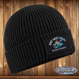 May include: Black ribbed knit beanie with a rectangular patch featuring a cartoon duck wearing a red hat and smoking a pipe. The patch reads "DAT HEET MOIN" and "MOIN MOIN IS GESABBEL!" in white text. The beanie is set against a wooden background.