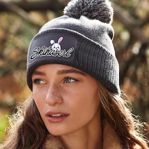 Könnte beinhalten: Graue Strickmütze mit Bommel und einem schwarzen Patch mit einem weißen Hasen und dem Text "Skihaserl" auf der Vorderseite.